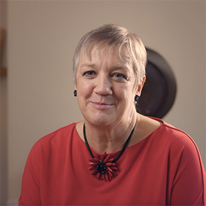 Portrait of Sue Nelson smiling, wearing a red top and flower necklace